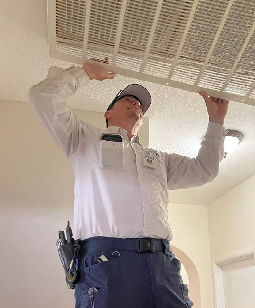 YES! technician wearing a cap and uniform inspecting an air vent while holding the vent cover overhead, standing on a ladder.