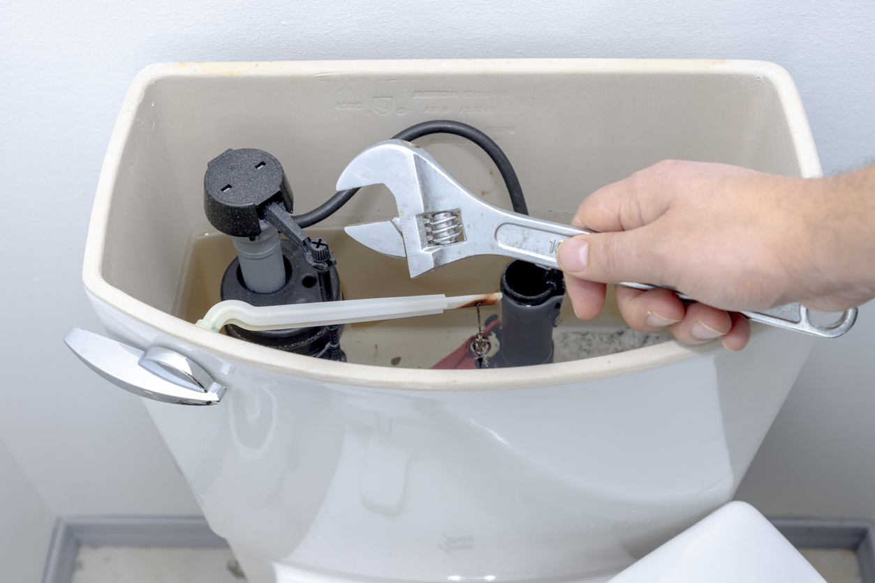 A man holding an adjustable wrench with an open tank exposing the interior valve and lever, about to conduct toilet maintenance checks.