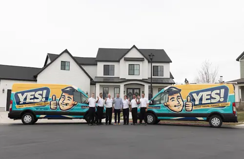 The YES! team of HVAC and plumbing professionals stands in front of two branded vans outside a home, smiling.