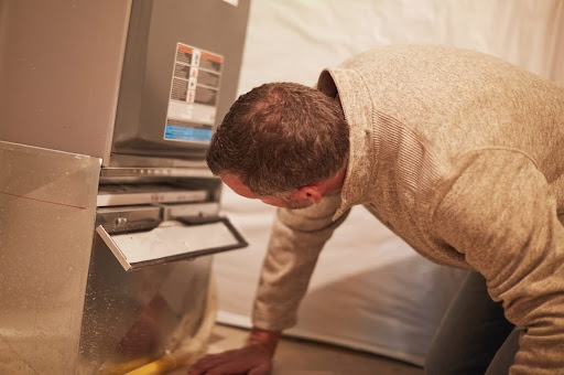 A man kneeling on a basement floor, looking under the open access panel of a home furnace or heater.