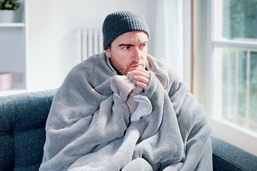 Man dressed warm in a blanket and wearing a beanie, sitting on a couch and looking cold, with a broken radiator in the background.