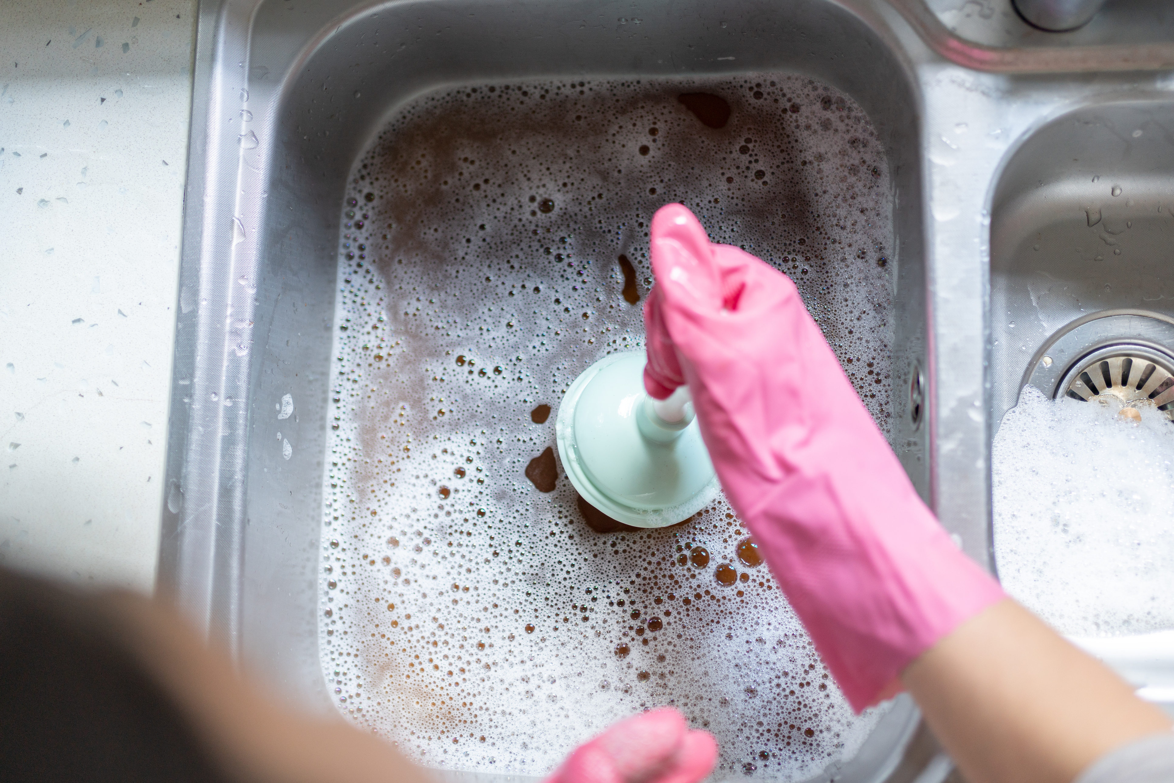 Woman Using Plunger In Blocked Kitchen Sink To Unclog Drain.